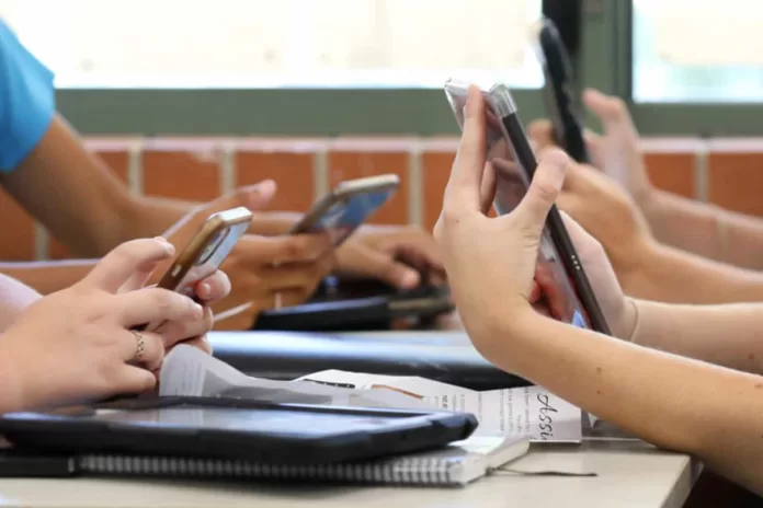 Mãos de crianças em suas carteiras segurando seus celulares.