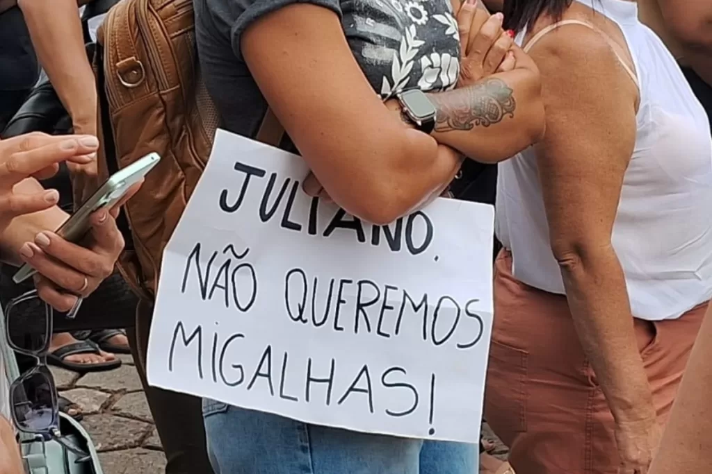 Mulher com os braços cruzados segura um cartaz escrito "Juliano, não queremos migalhas!"