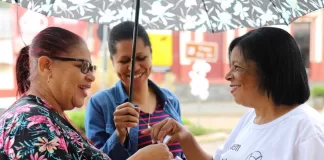 Uma agente de saúde, vestida com uma camiseta branca, entrega papéis da campanha para duas mulheres na rua. Uma delas, usa uma sombrinha para protegê-las da chuva.