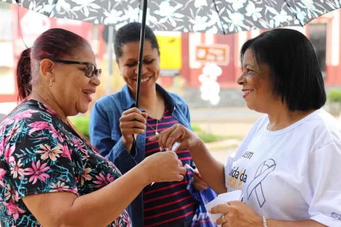 Uma agente de saúde, vestida com uma camiseta branca, entrega papéis da campanha para duas mulheres na rua. Uma delas, usa uma sombrinha para protegê-las da chuva.