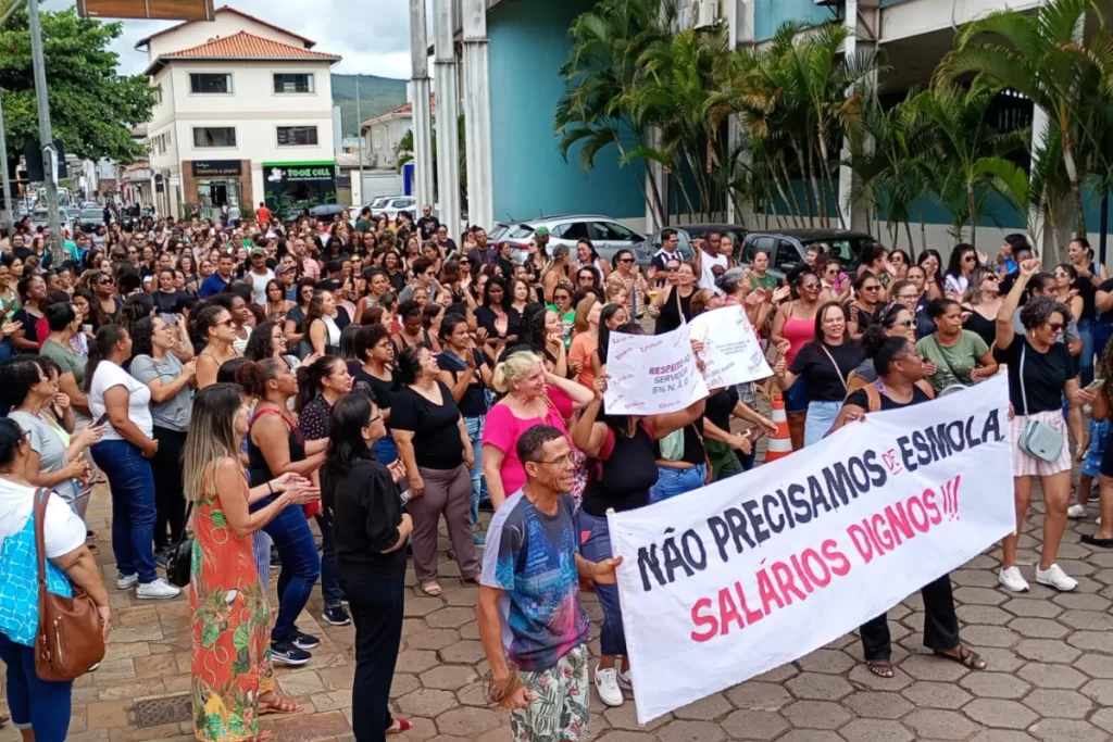 Manifestantes caminham pela rua atrás da prefeitura com cartazes.