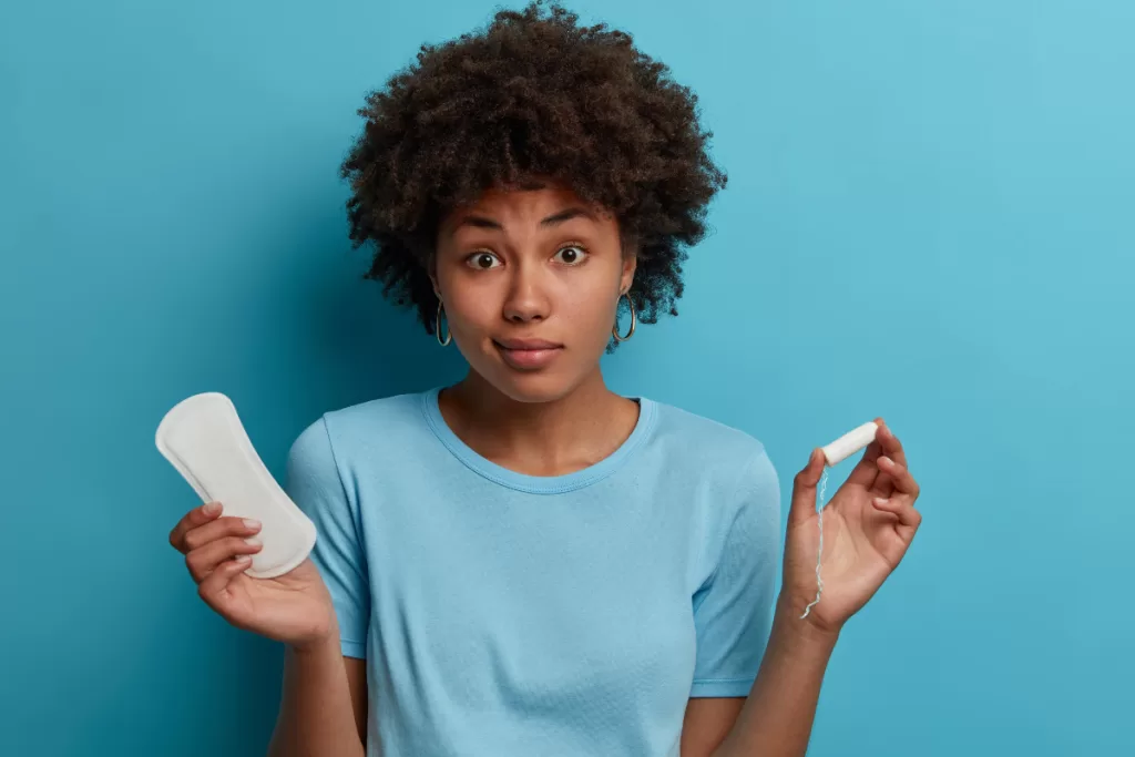 Moça de blusa azul segurando um absorvente interno numa mão e outro absorvente sem abas em outra. Ela olha para o público com uma cara de dúvida.